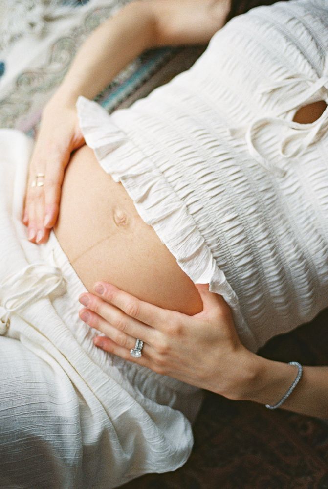 A pregnant woman in a white dress touches her belly while wearing a ring and bracelet.