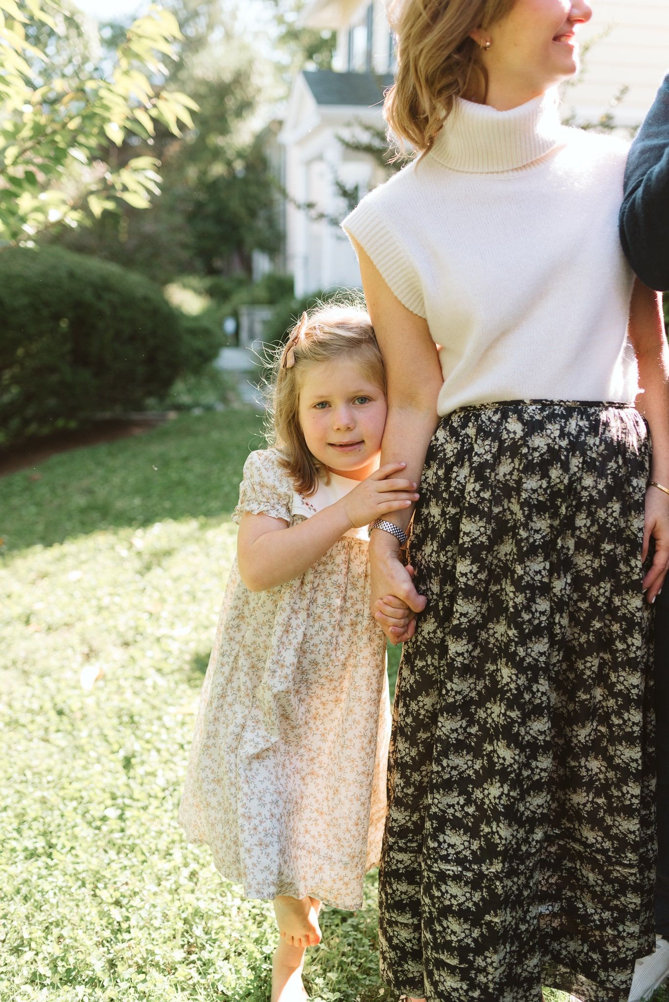 A mother and young child stand together in a garden wearing coordinating floral and white outfits.