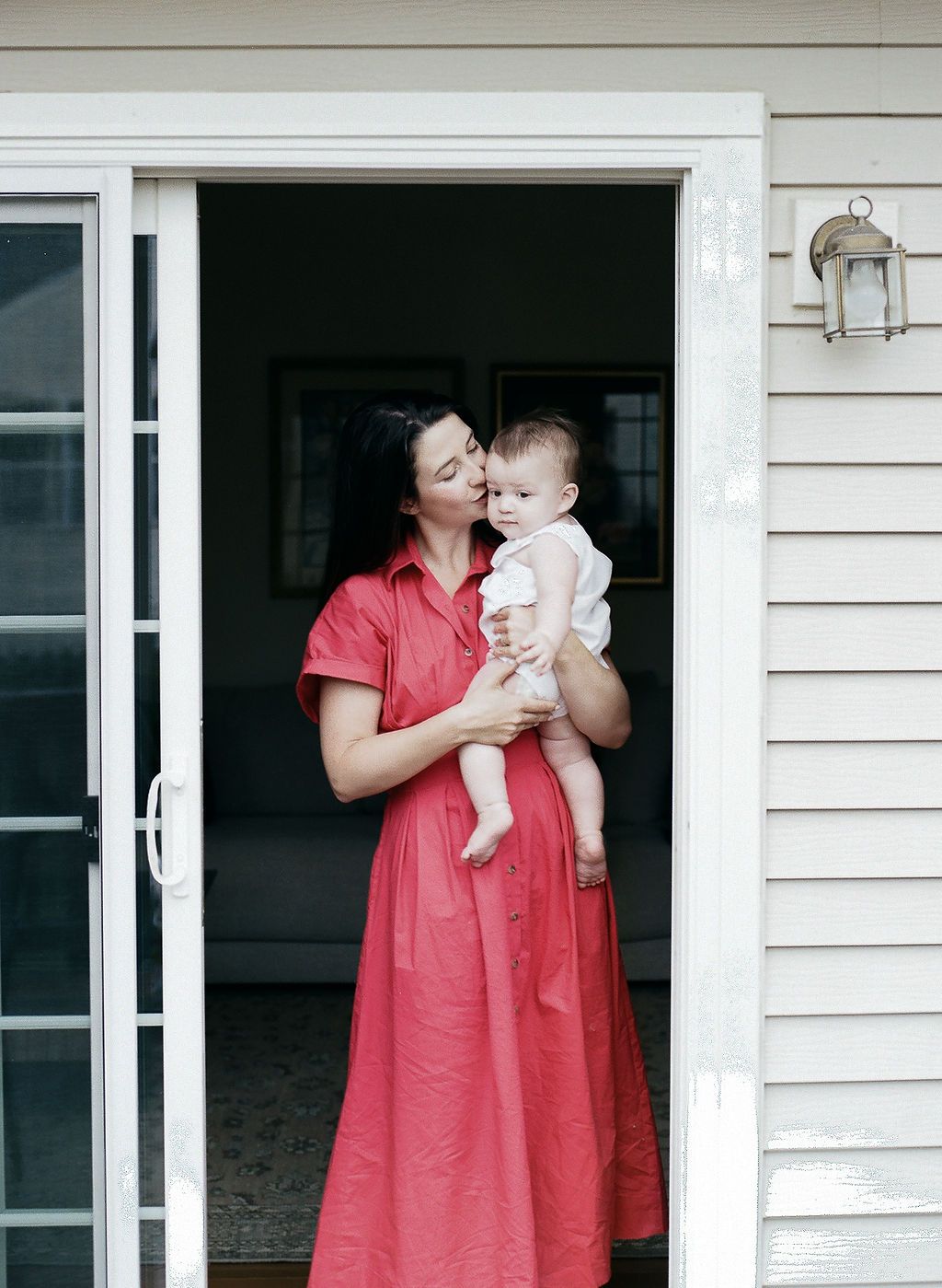 A series of outdoor portraits showing intimate moments between a parent in red dress and a baby beside a white house.
