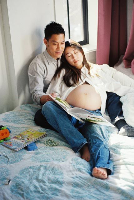 Pregnant Asian couple sitting on bed reading a book together, woman leaning on man's shoulder.