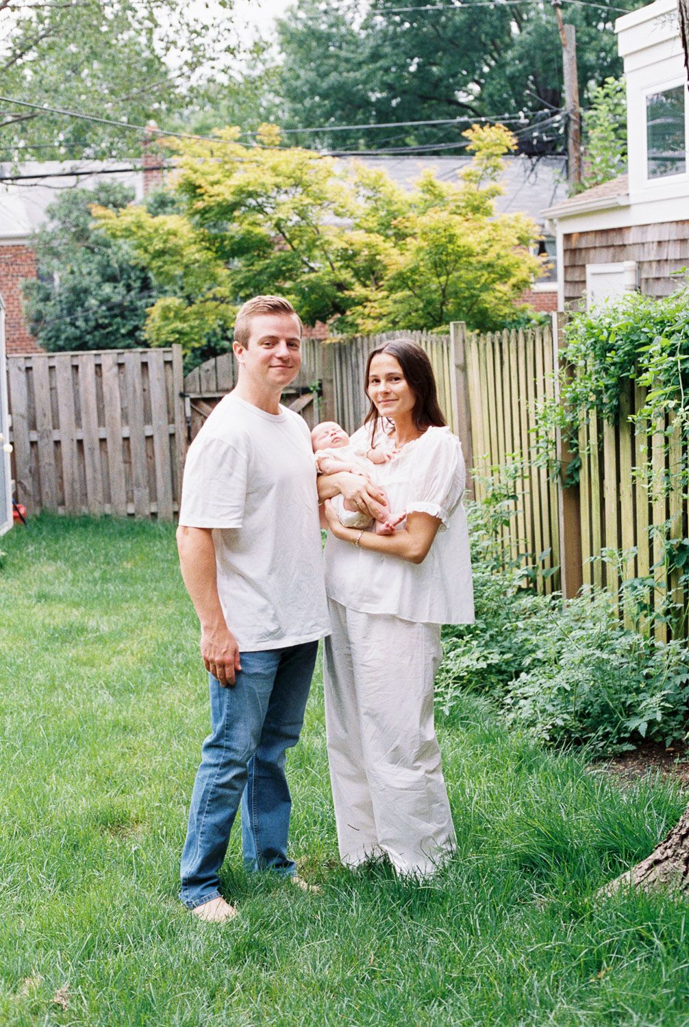 A couple stands in a backyard holding their newborn baby, dressed in casual white outfits, surrounded by green trees and a wooden fence.