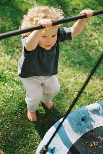 Curly-haired toddler standing barefoot on grass, holding a metal bar near a trampoline outdoors.