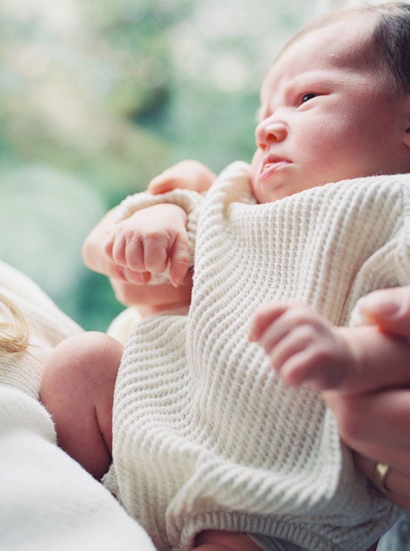 A sleeping newborn baby wearing a cream knit sweater stretches their tiny hands against a soft blurred background.