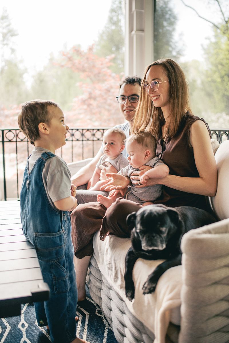 Happy family with two babies and a toddler sitting on a porch sofa with a black dog, sharing a joyful moment.