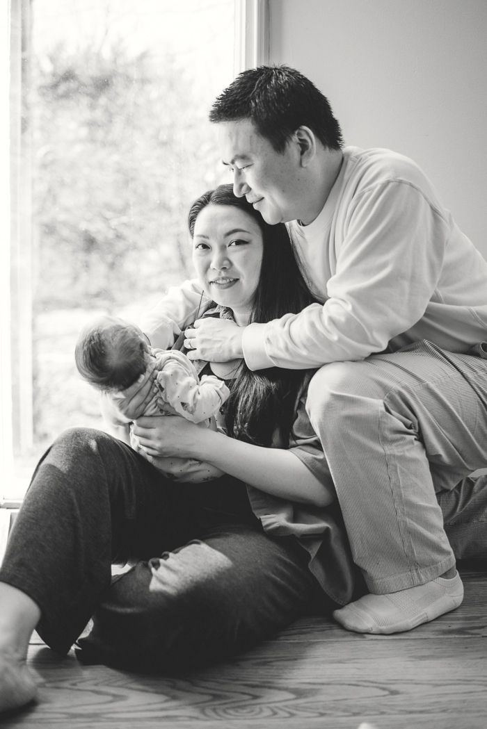 Black and white photo of Asian parents sitting on floor by window, mother holding newborn baby, father embracing them both.