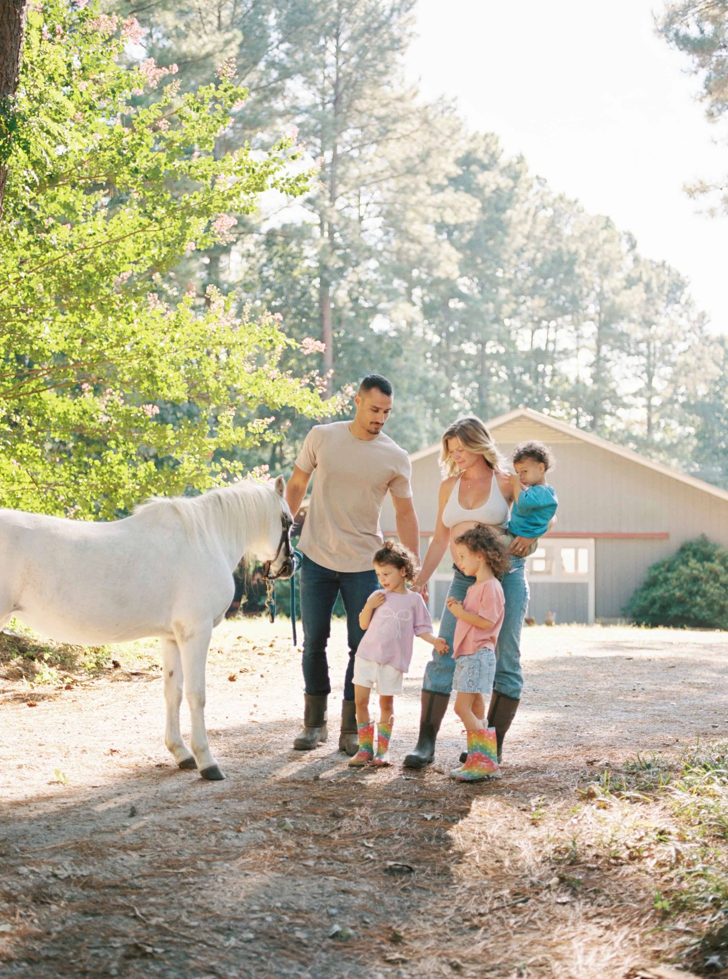 Family walking together with white horse on tree-lined path near barn.