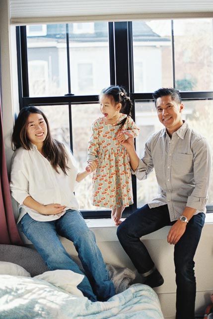Happy Asian family of three posing by a large window, parents sitting with toddler daughter in floral dress.