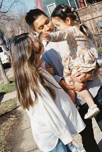 A couple smiles at their young daughter being held by the man on a sunny sidewalk.