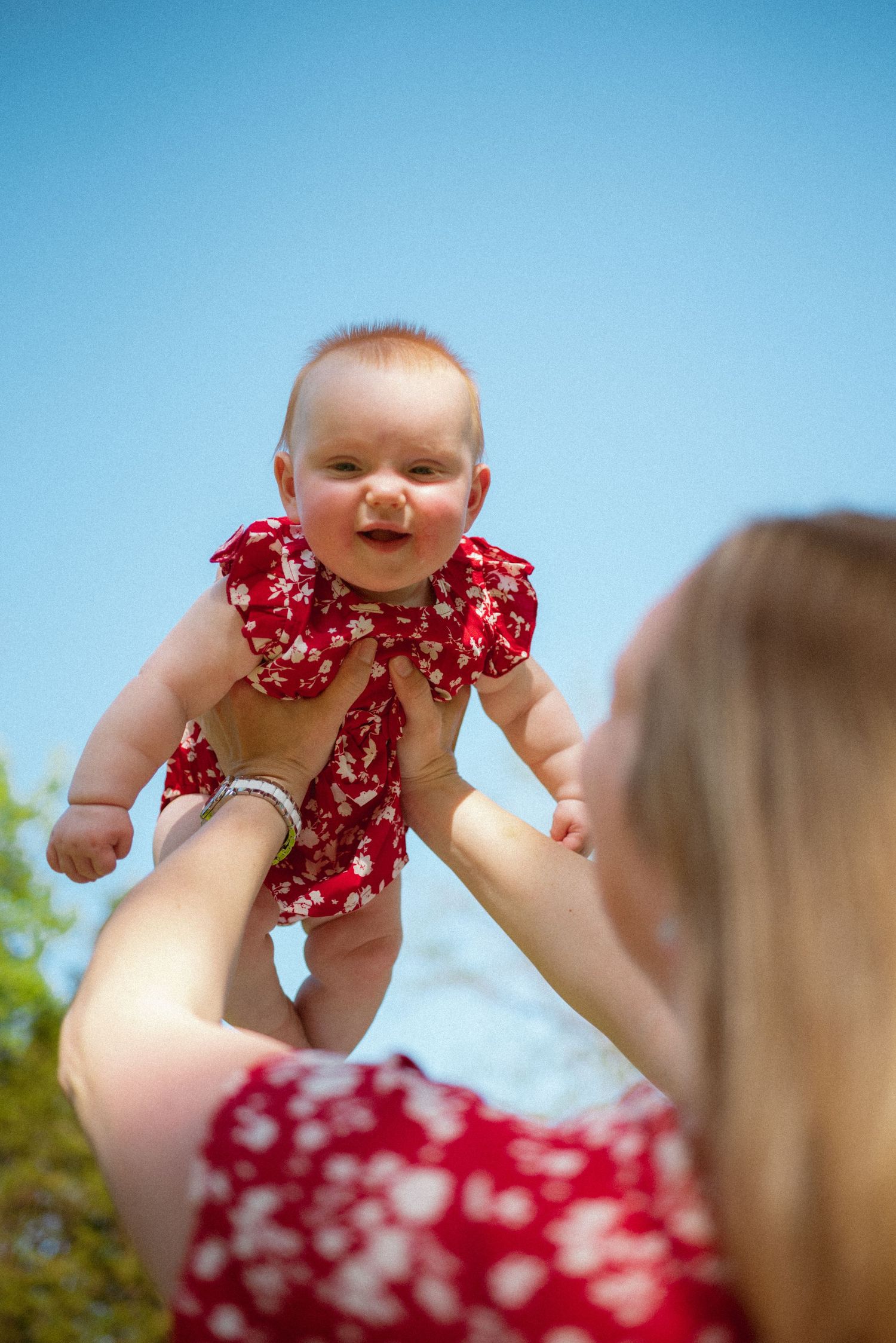 An infant wearing a red floral outfit is lifted high into the air against a bright blue summer sky.