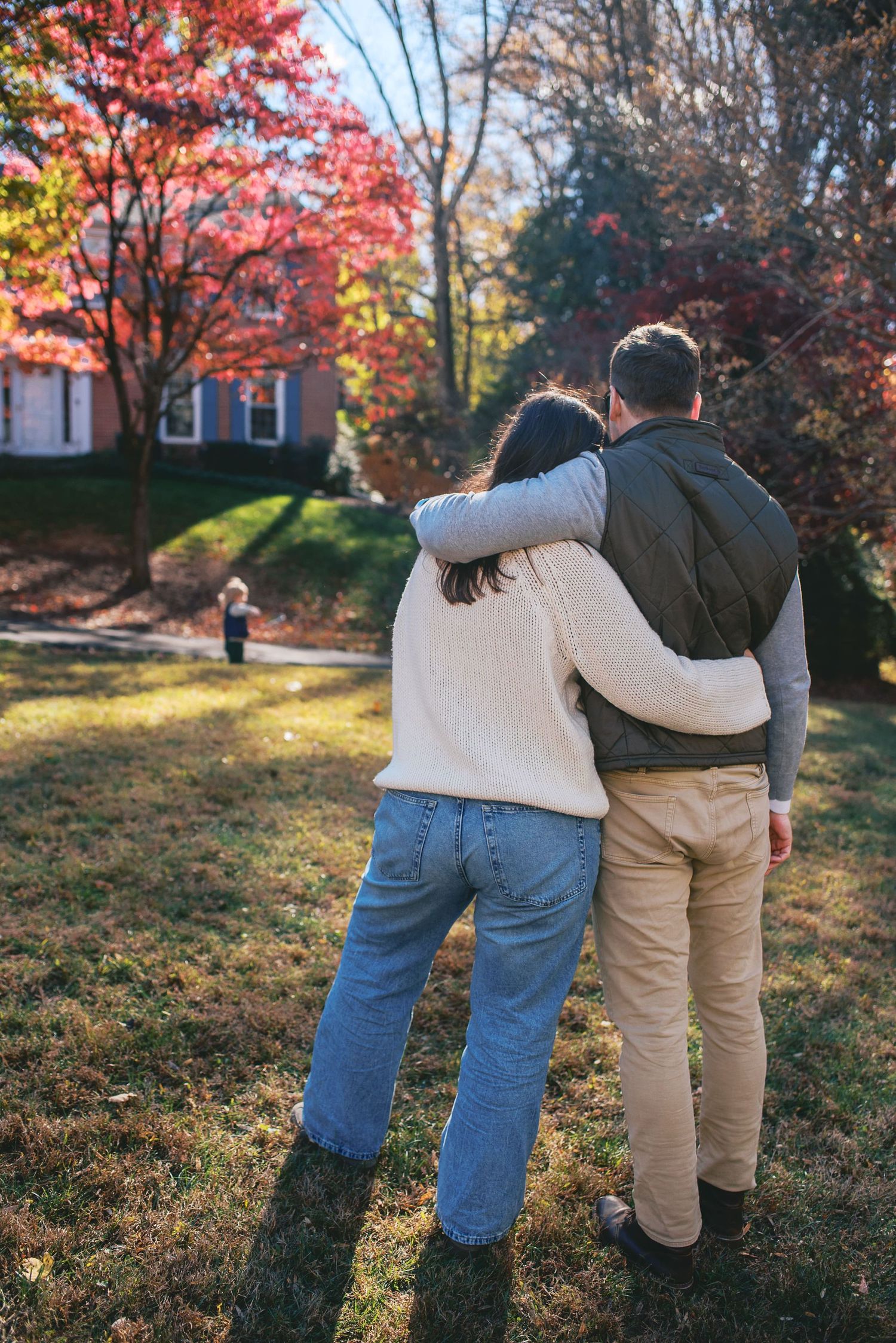 A couple embraces while walking through a scenic autumn landscape with vibrant red maple trees in the background.
