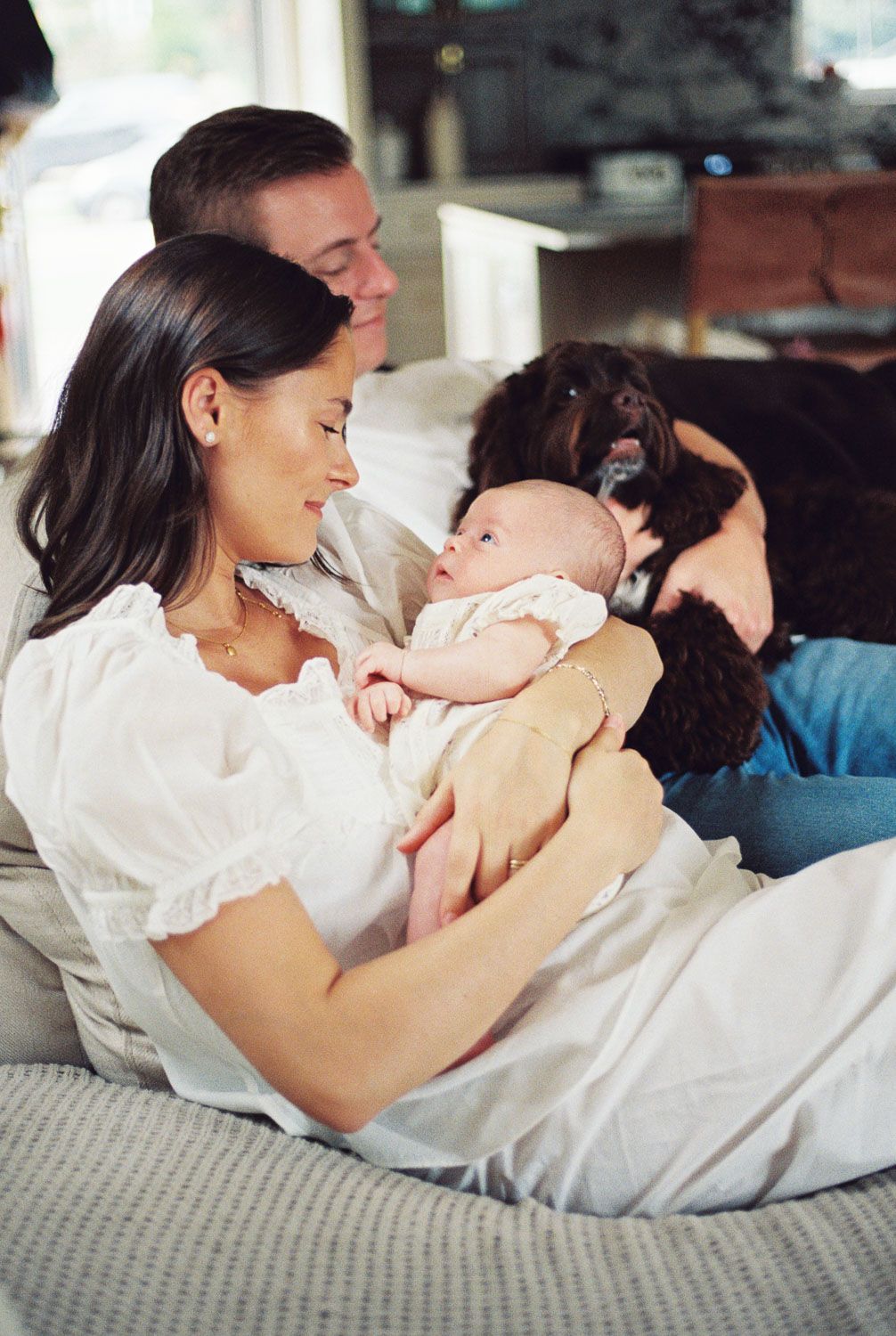 A young family sits on a couch, mother holding newborn baby, father and black dog beside them, in a bright home.