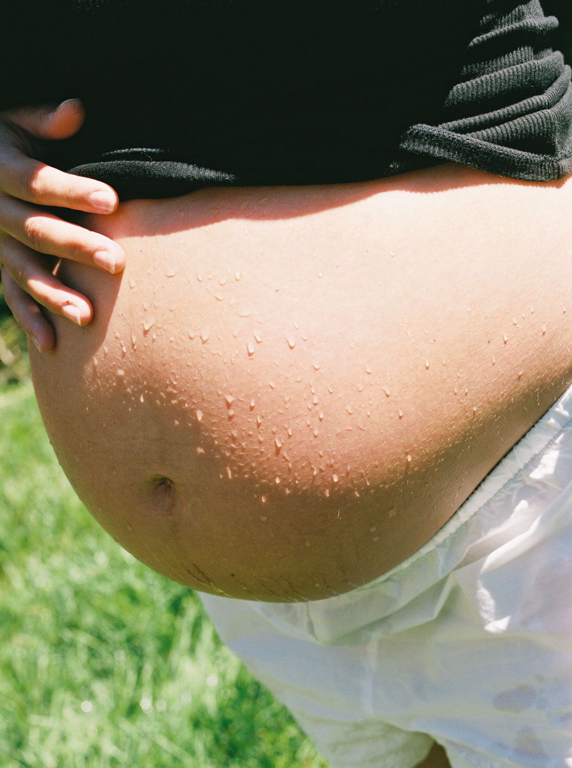 Close-up of a pregnant woman's bare wet belly outdoors with green grass in the background.