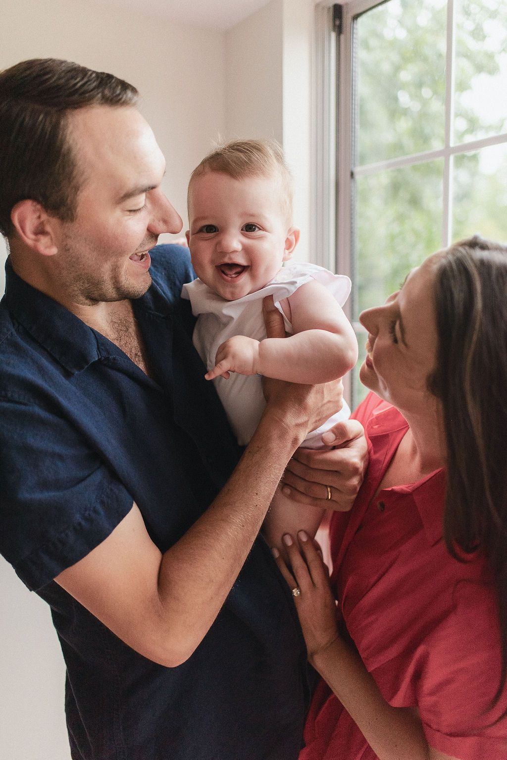 A joyful family moment captured with a baby being lifted and cuddled indoors by window light.
