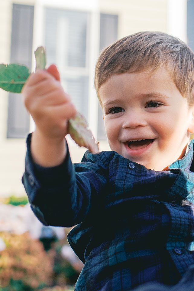 A young child in a navy blue jacket raises their arms joyfully while playing outdoors on a sunny day.