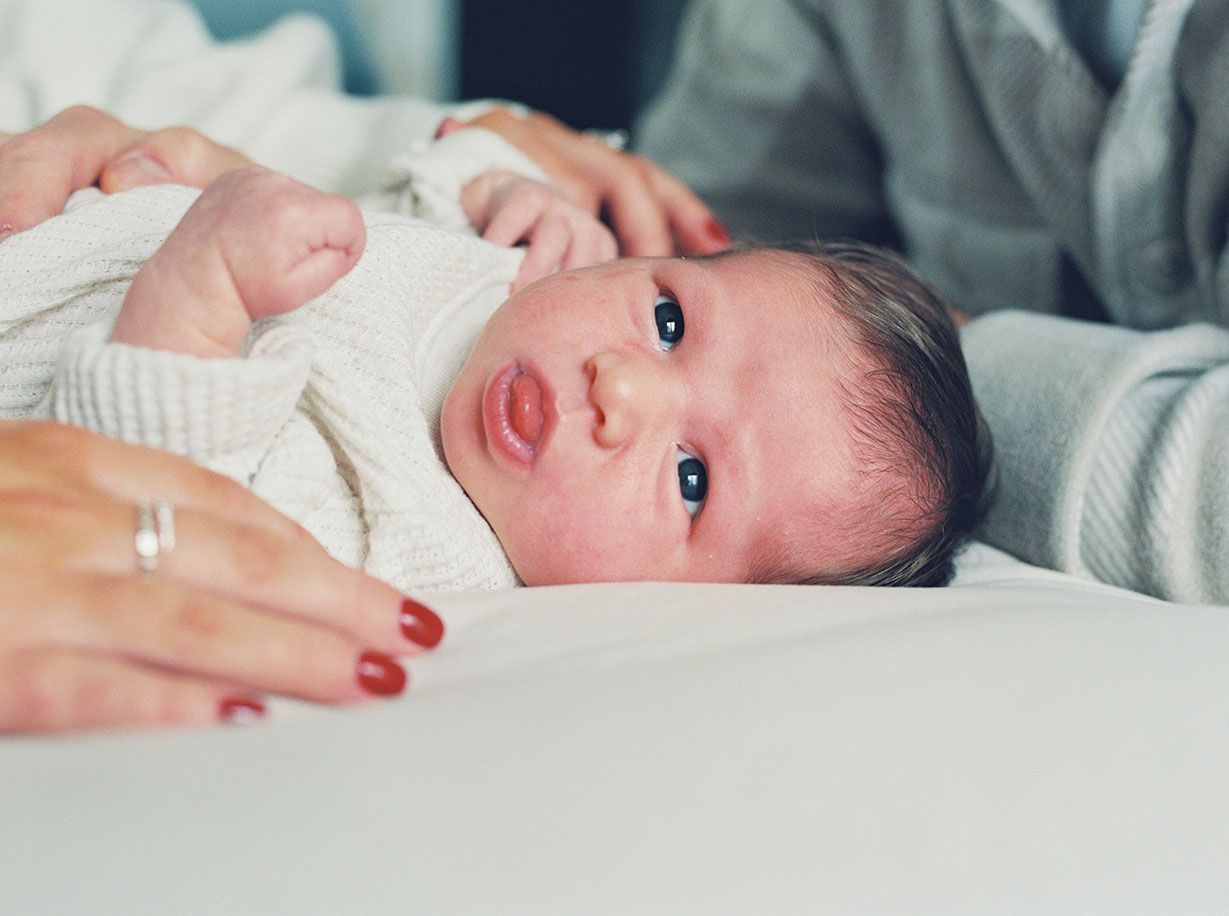 Close up of a newborn baby with rosy cheeks wearing a white outfit.