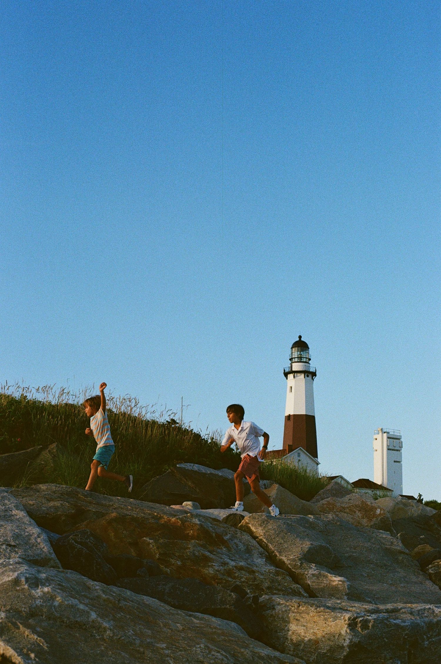 Two people hiking on rocky terrain near Montauk Lighthouse with a clear blue sky in the background.