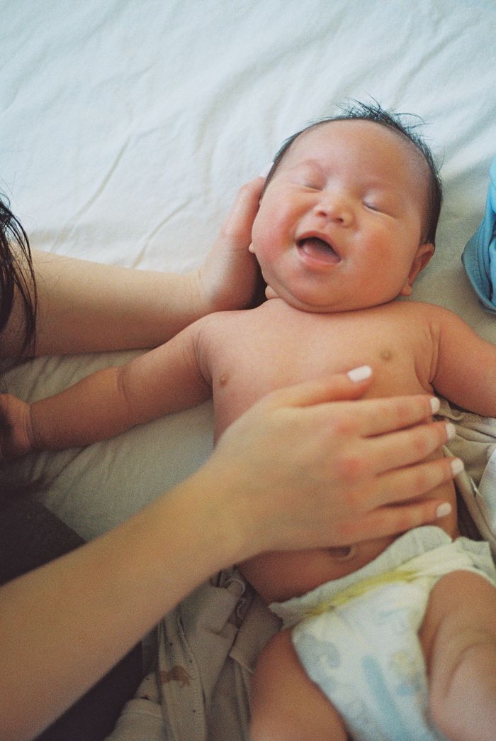 A newborn baby smiling while lying on a white bed, being gently held by a parent's hands.