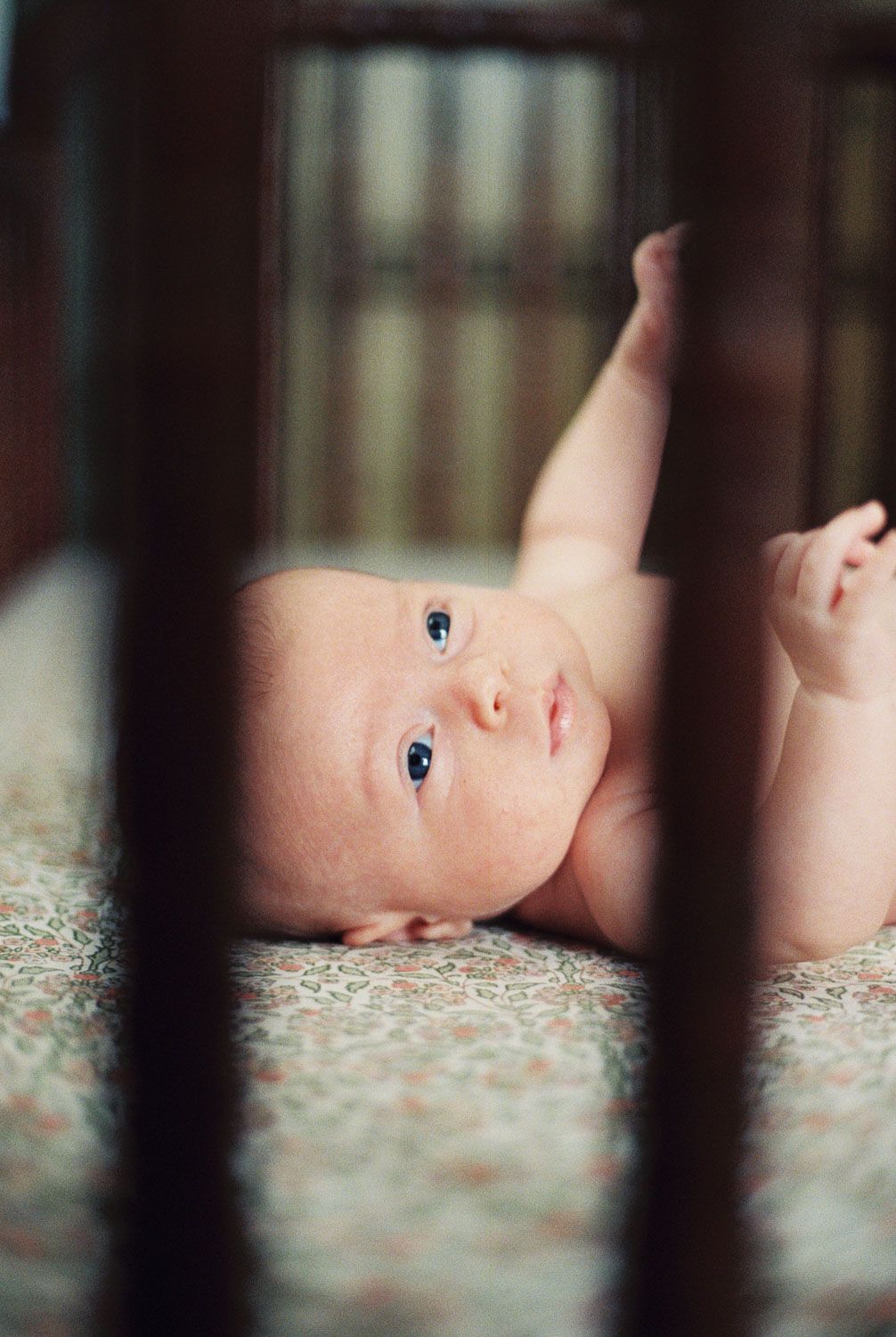 Newborn baby lying on back in a wooden crib, looking up with blue eyes, arms raised, on a floral mattress.