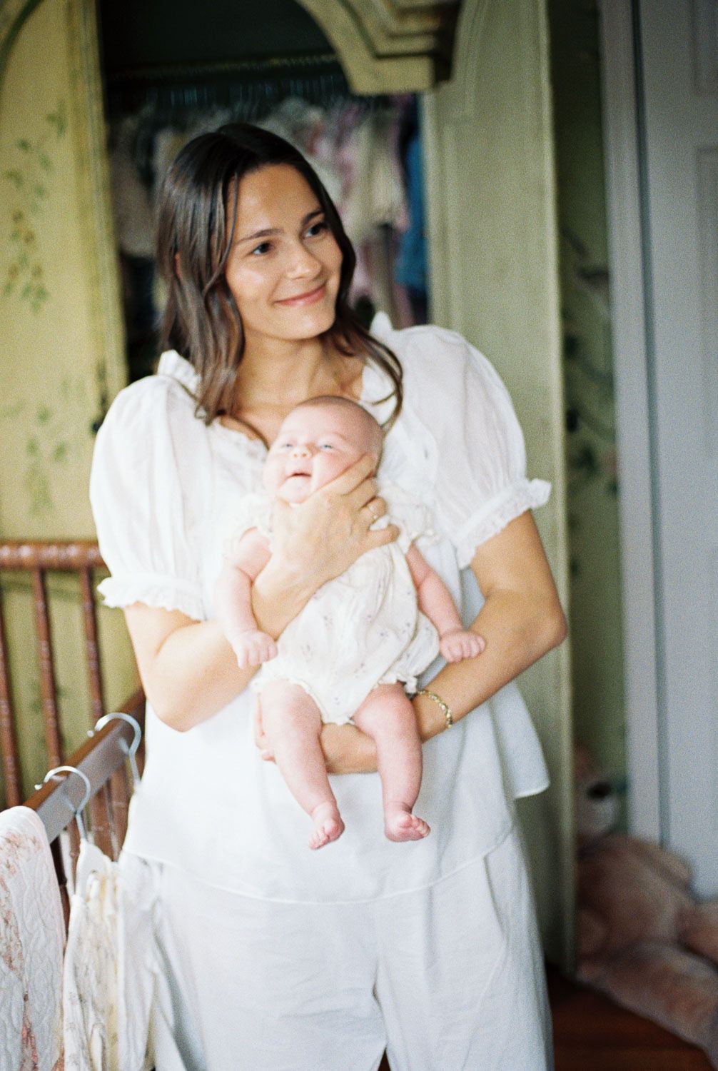 A smiling woman in white holds a happy baby dressed in white, standing in a softly decorated room.