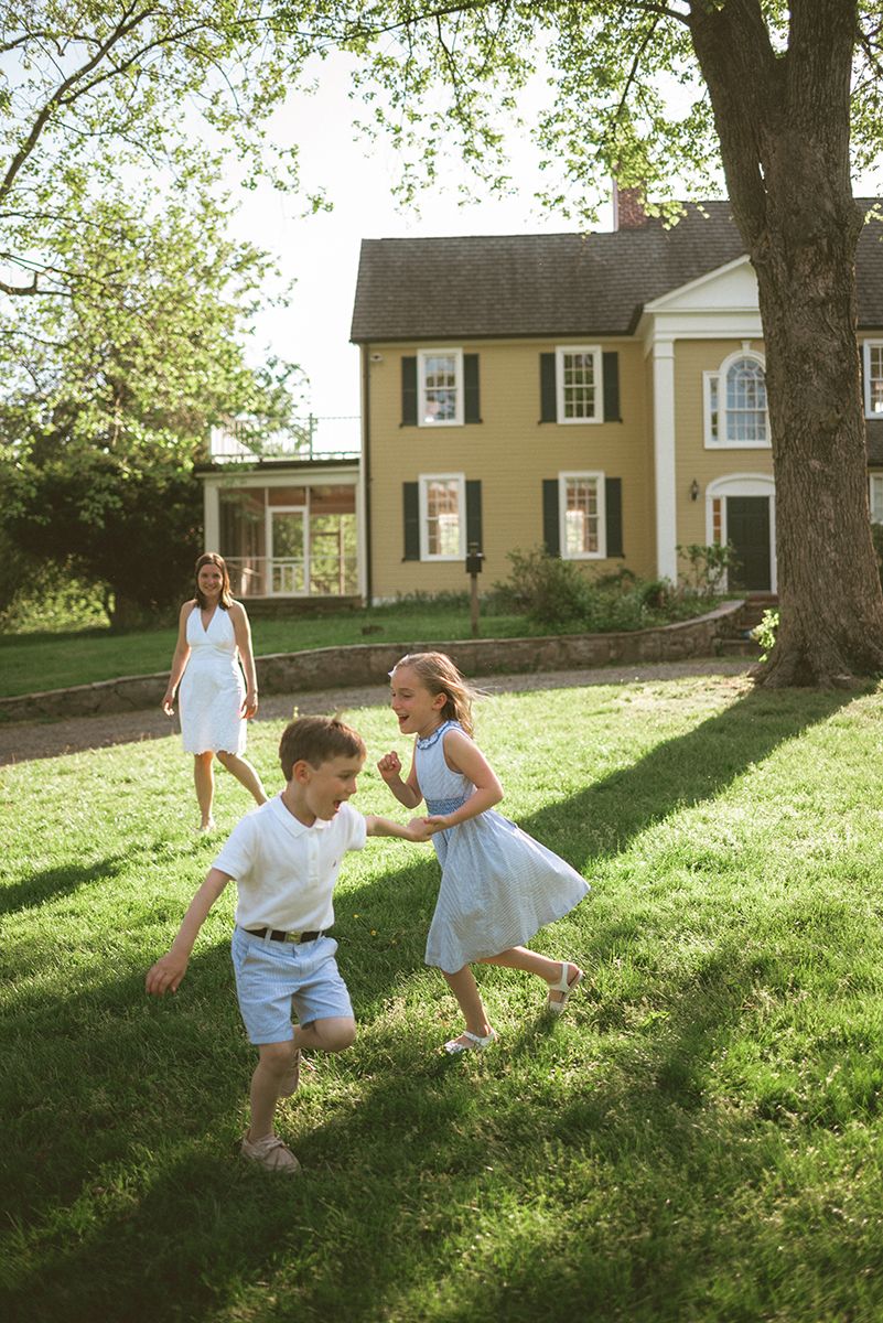 Children play and run in a sunlit yard in front of a yellow colonial-style house with white trim.