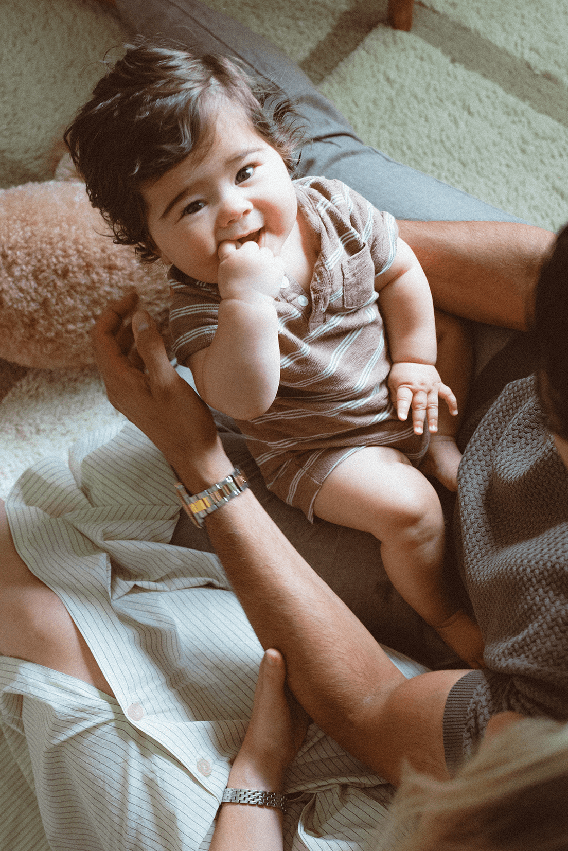 Happy baby in gray romper sits on white blanket with caregiver, making a sweet joyful expression.