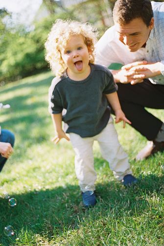 Curly-haired toddler sticking out tongue outdoors on grass with smiling man crouching behind, playing with bubbles.