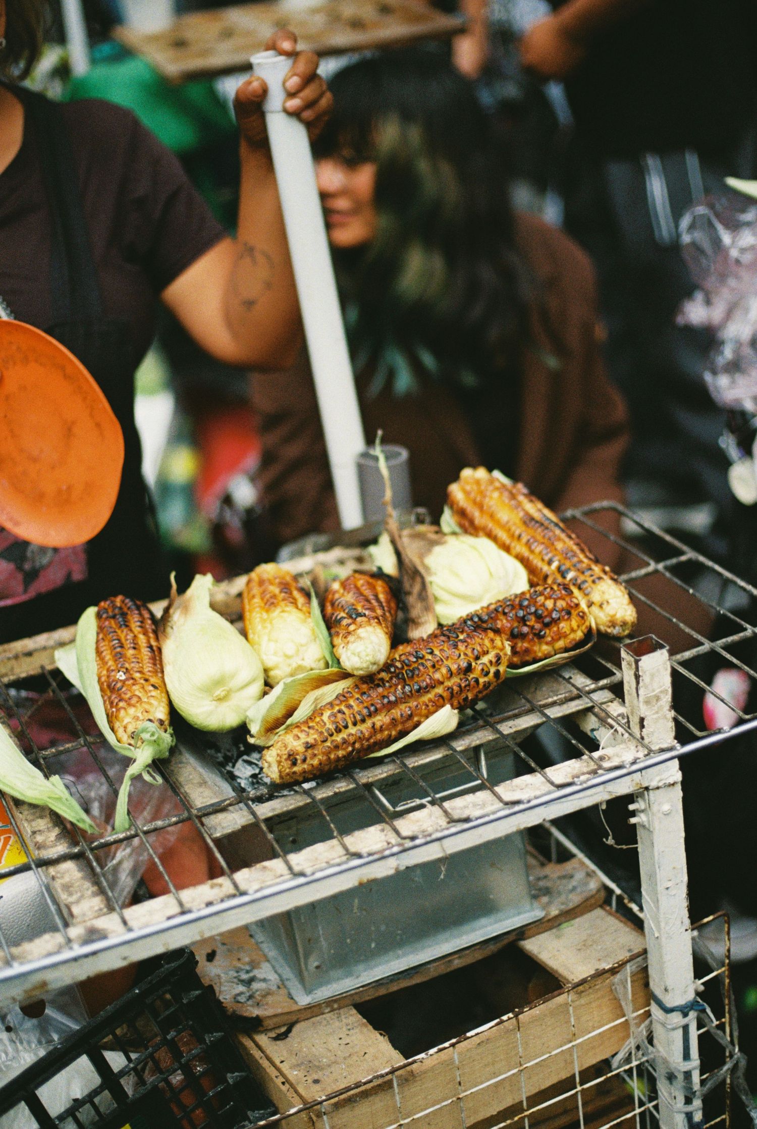 Grilled corn and vegetables cooking on a metal grill at an outdoor food market or street vendor stall.
