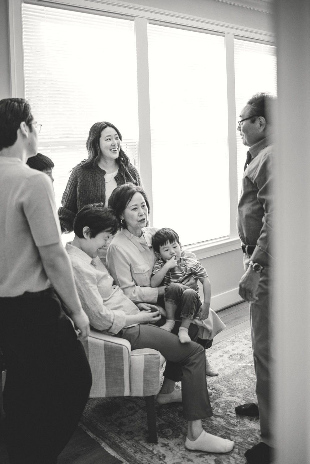 Black and white family portrait with multigenerational Asian family gathered in bright living room, grandmother holding toddler.