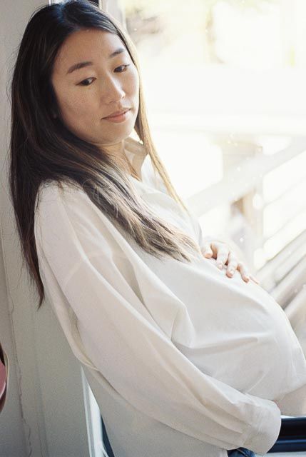 Pregnant Asian woman in white blouse cradling her baby bump while leaning against a window in soft natural light.