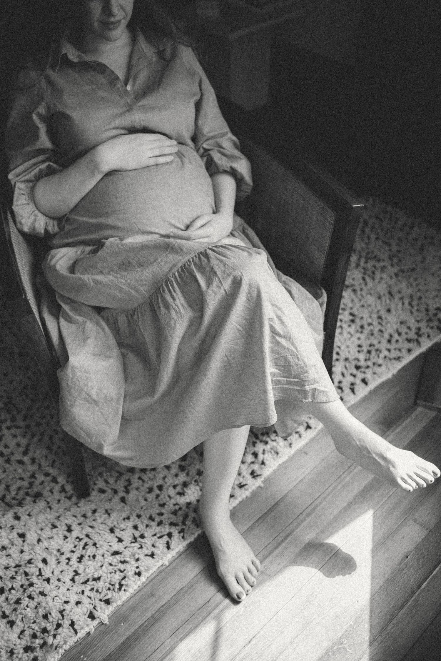 Black and white photograph showing someone in a light dress sitting on a chair, resting their bare feet on wooden flooring.
