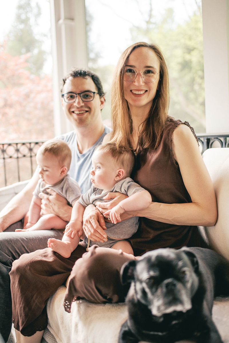 Family portrait of two parents with twin babies and a black dog sitting on a couch in a bright living room.