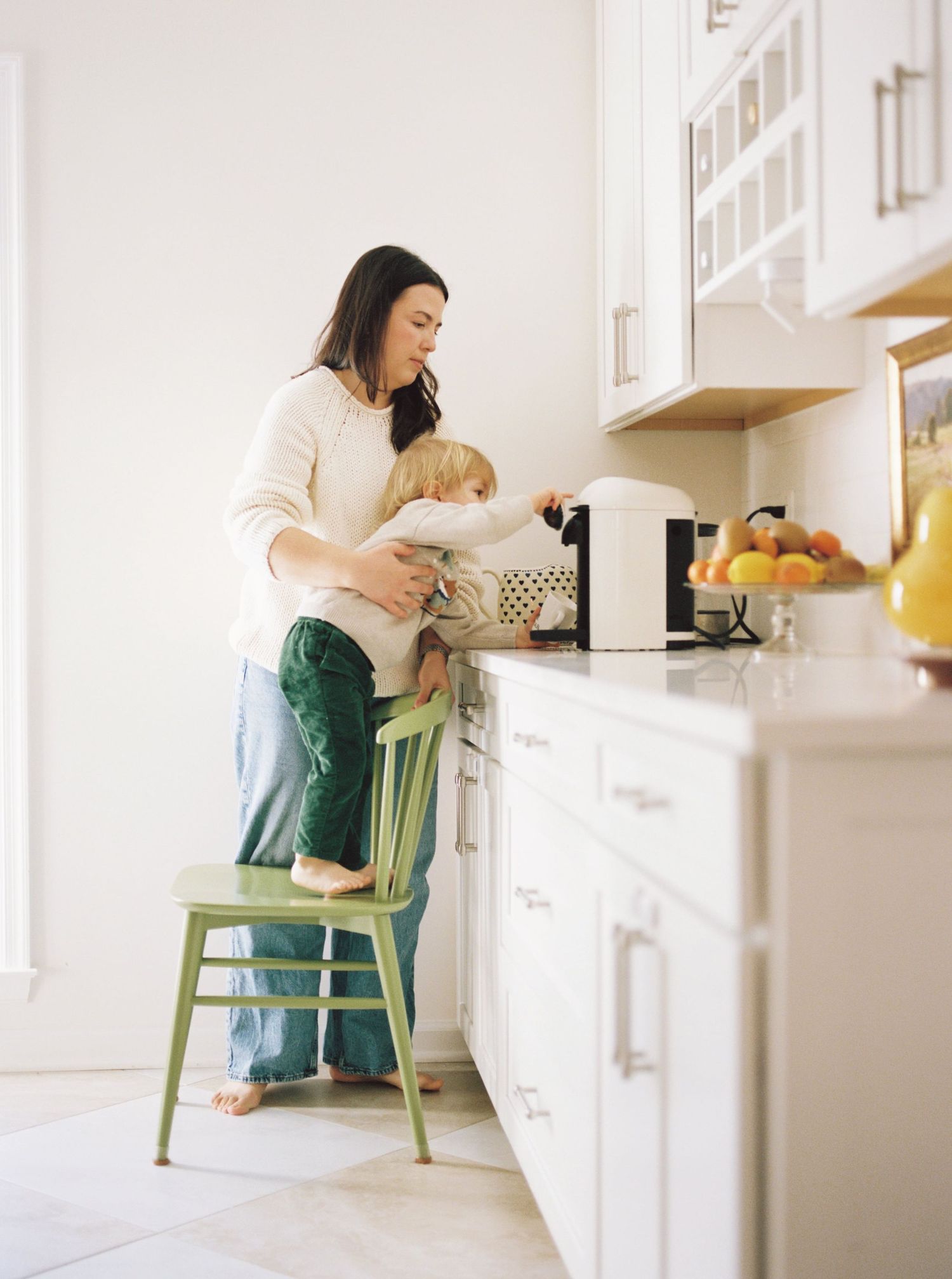 A woman in green overalls stands at a white kitchen counter preparing food while her child sits nearby on a stool.