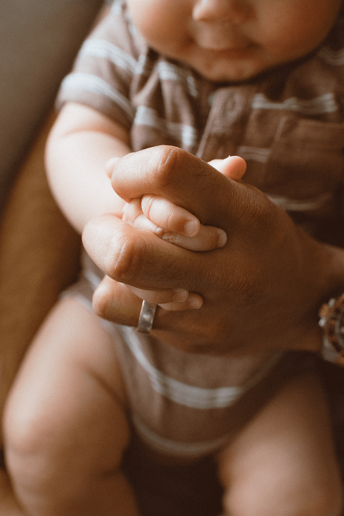 Close up view of adult and baby hands clasped together, with rings visible in warm lighting.