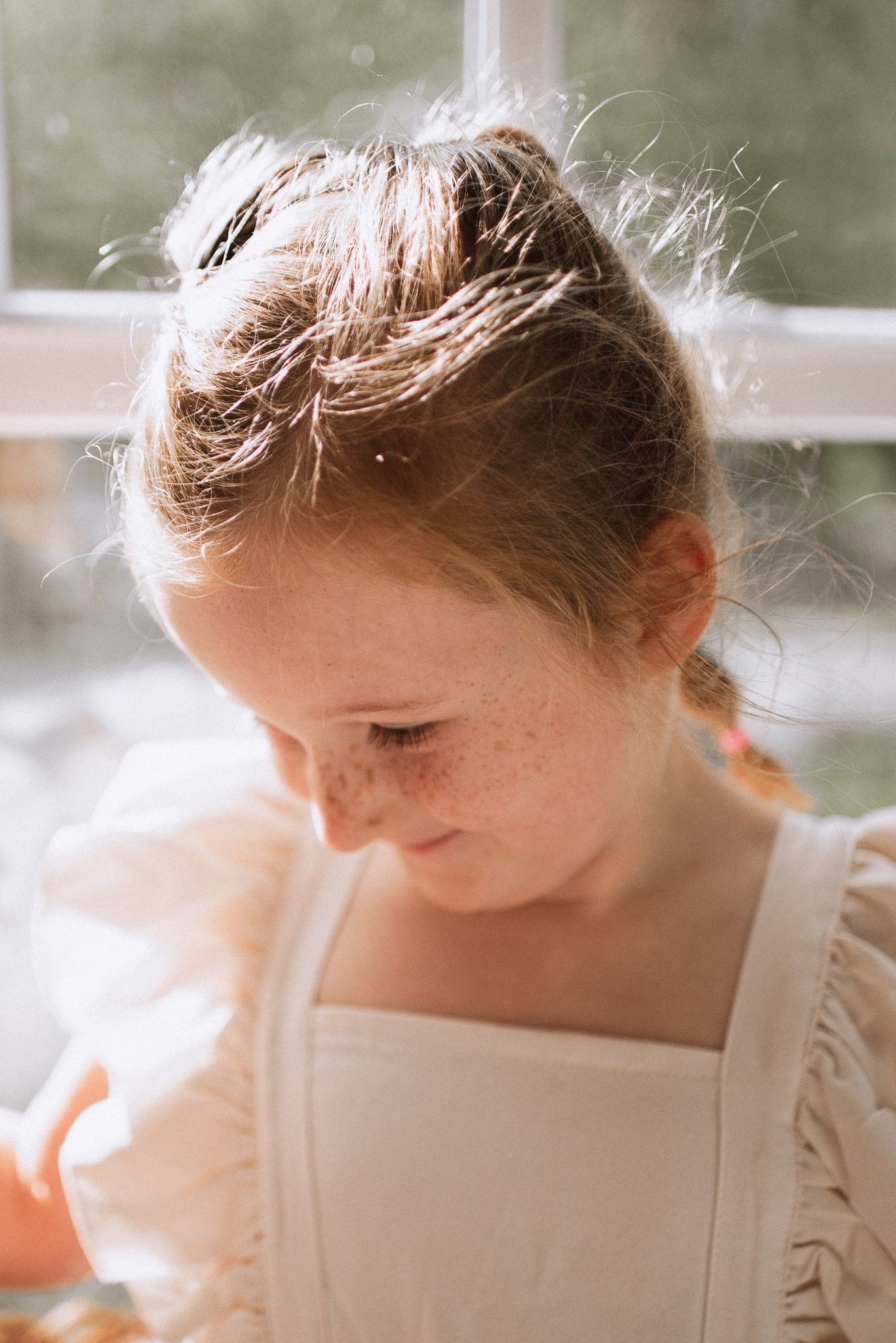 Soft sunlit portrait shows a child with freckles and wispy hair in a white dress looking down with a gentle smile.
