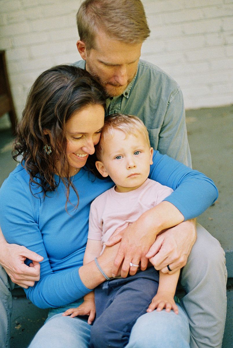 Happy family portrait with mother, father, and toddler in pink shirt sitting together against white brick wall.