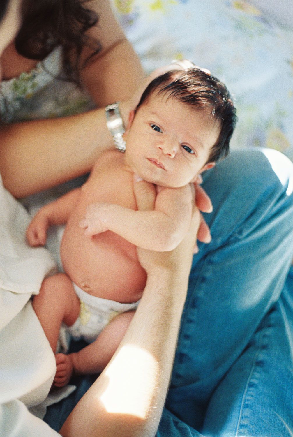Newborn baby with dark hair and wide eyes, wearing a diaper, is cradled in a parent's arms in warm, soft sunlight.