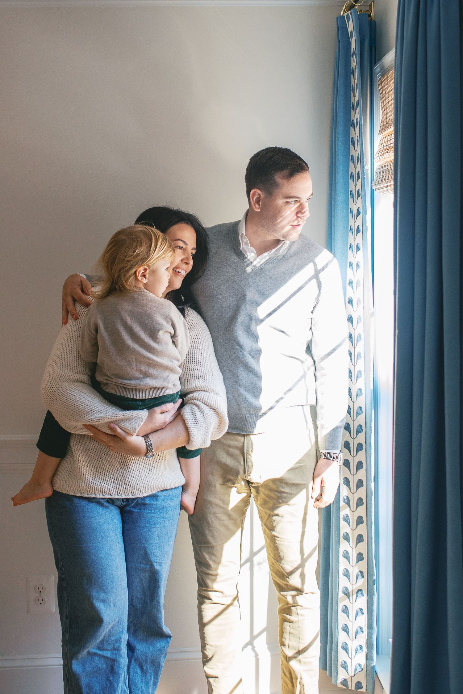A parent holds a small child near a window with blue curtains while looking outside together.