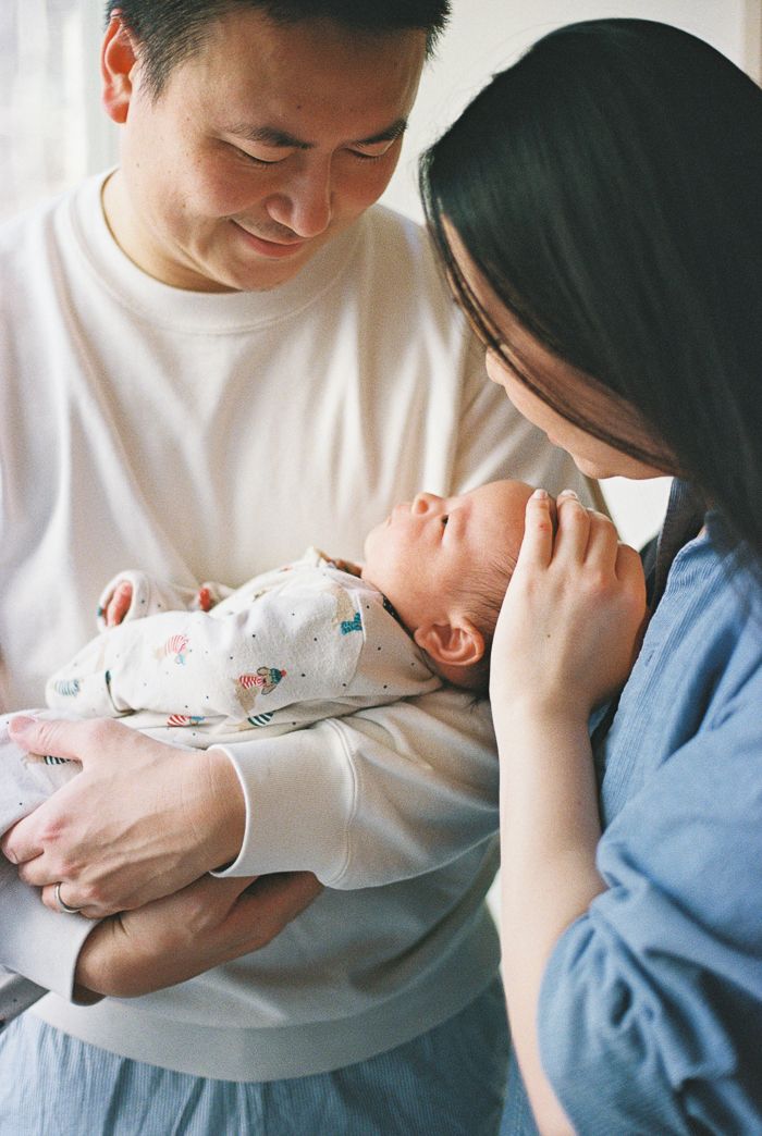 Asian parents lovingly gazing at their newborn baby, mother in blue dress cradling infant, father in white sweatshirt.