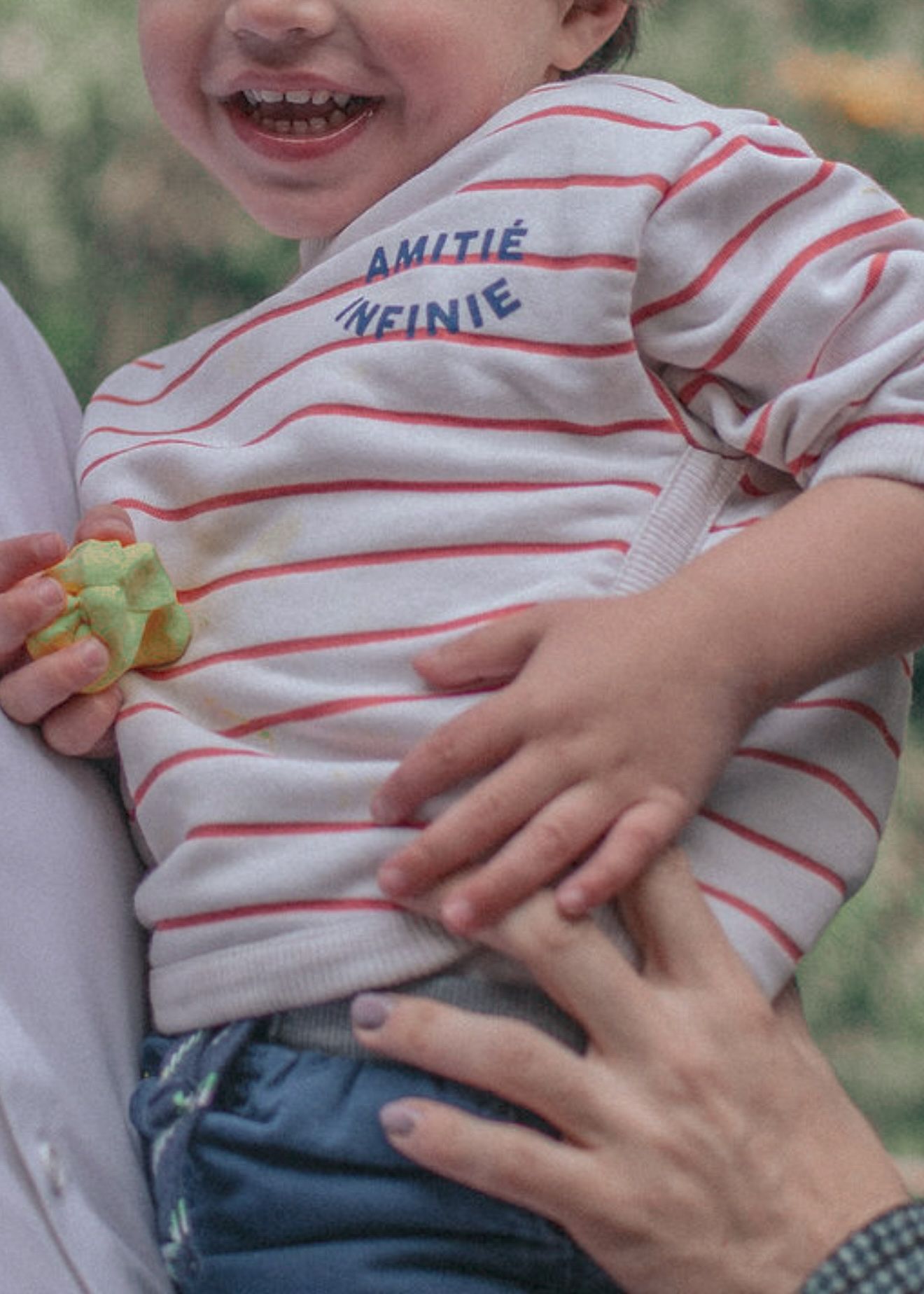 Small child in striped shirt holding pieces of fresh green fruit while being tickled by adult hands.