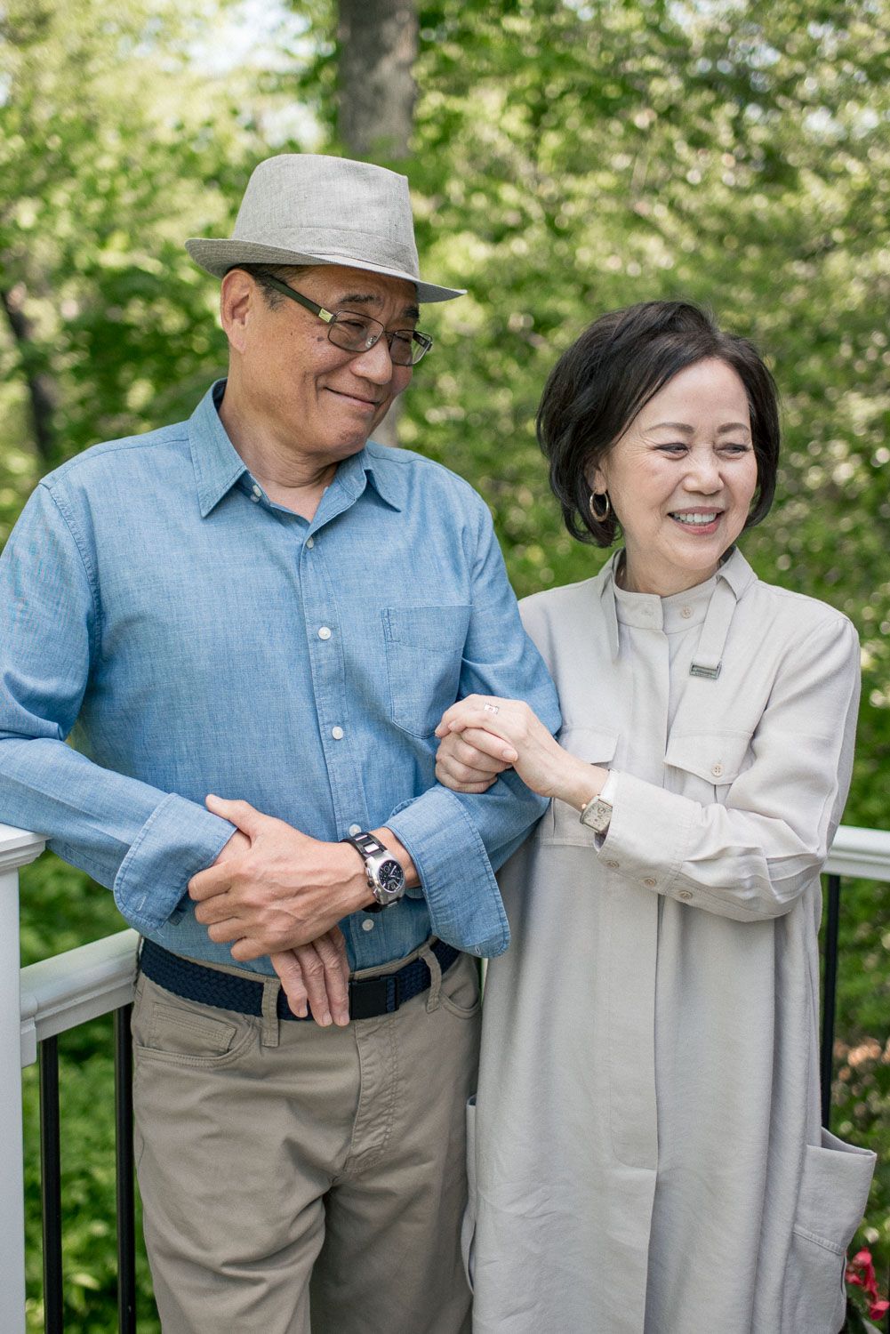 An older Asian couple smiles and holds hands on a white railing deck surrounded by lush green trees.