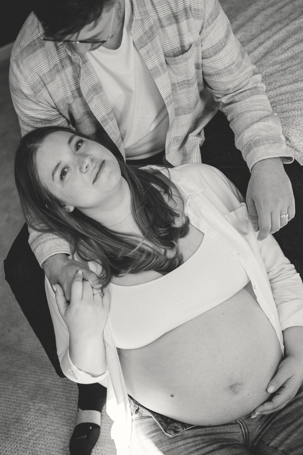 Black and white photo of a pregnant woman leaning against her partner, cradling her baby bump.