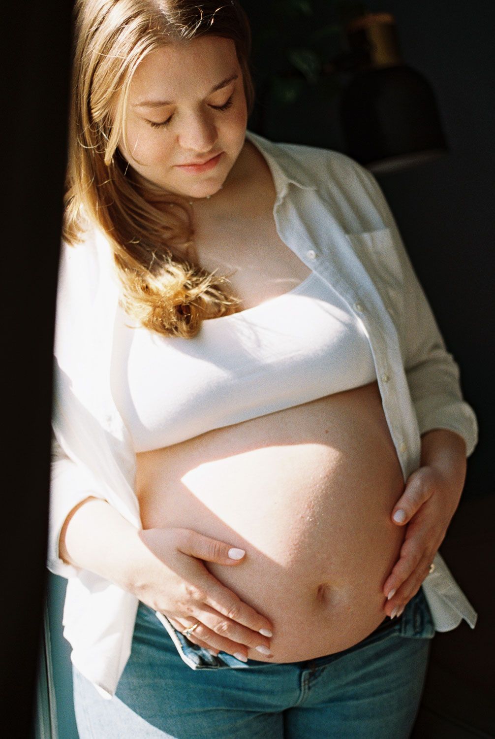 Pregnant woman in white shirt gently holding her bare baby bump in soft natural sunlight indoors.