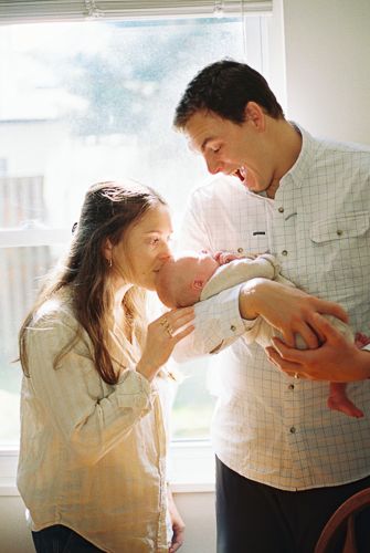 New parents smiling and bonding with their newborn baby by a bright window in a warm, intimate home setting.