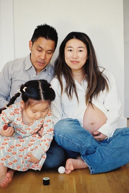 Asian family portrait with pregnant mother, father, and toddler daughter sitting on floor playing with toys.