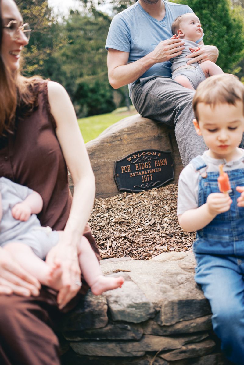 Family gathering at Fox Ridge Farm, established 1577, with parents holding babies near a stone wall outdoors.