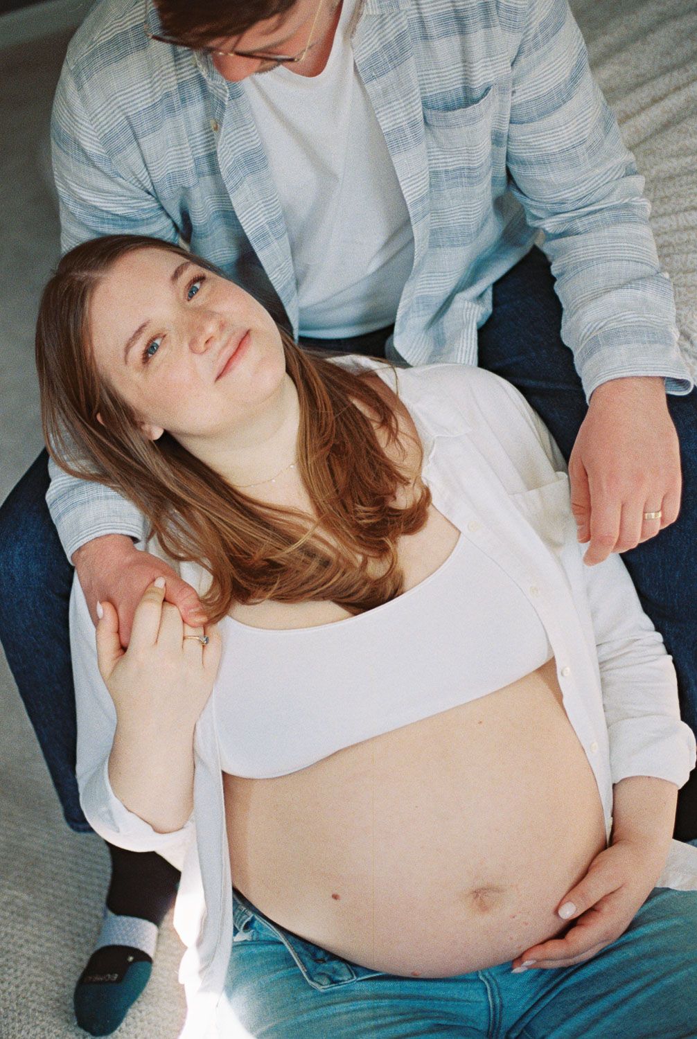 Pregnant woman sitting on floor leaning against partner, cradling bare baby bump in casual maternity photo.