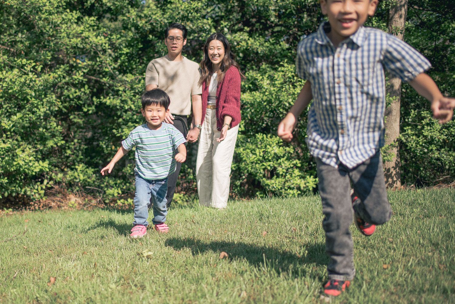 Asian family with two young boys running and playing on green grass in a sunny outdoor park setting.