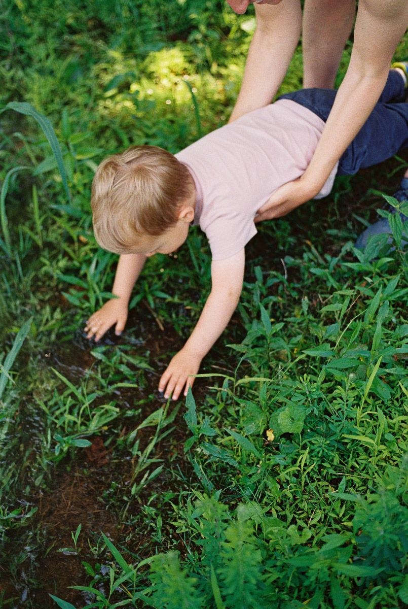 A toddler in a pink shirt leans toward a muddy garden patch while an adult holds them steady outdoors.
