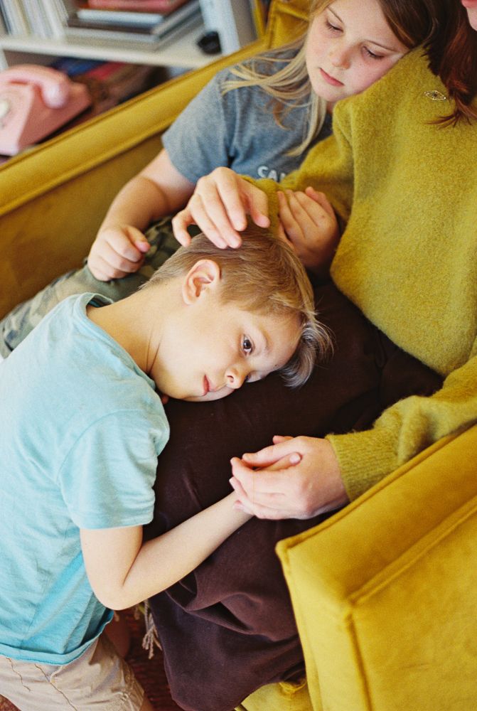 A child in a light blue shirt lies sleepily on a yellow couch while receiving a gentle head massage.