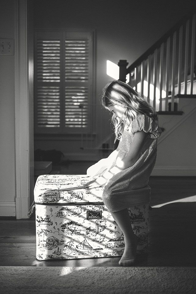A black and white photo of a small child exploring a decorative storage chest in natural window light.
